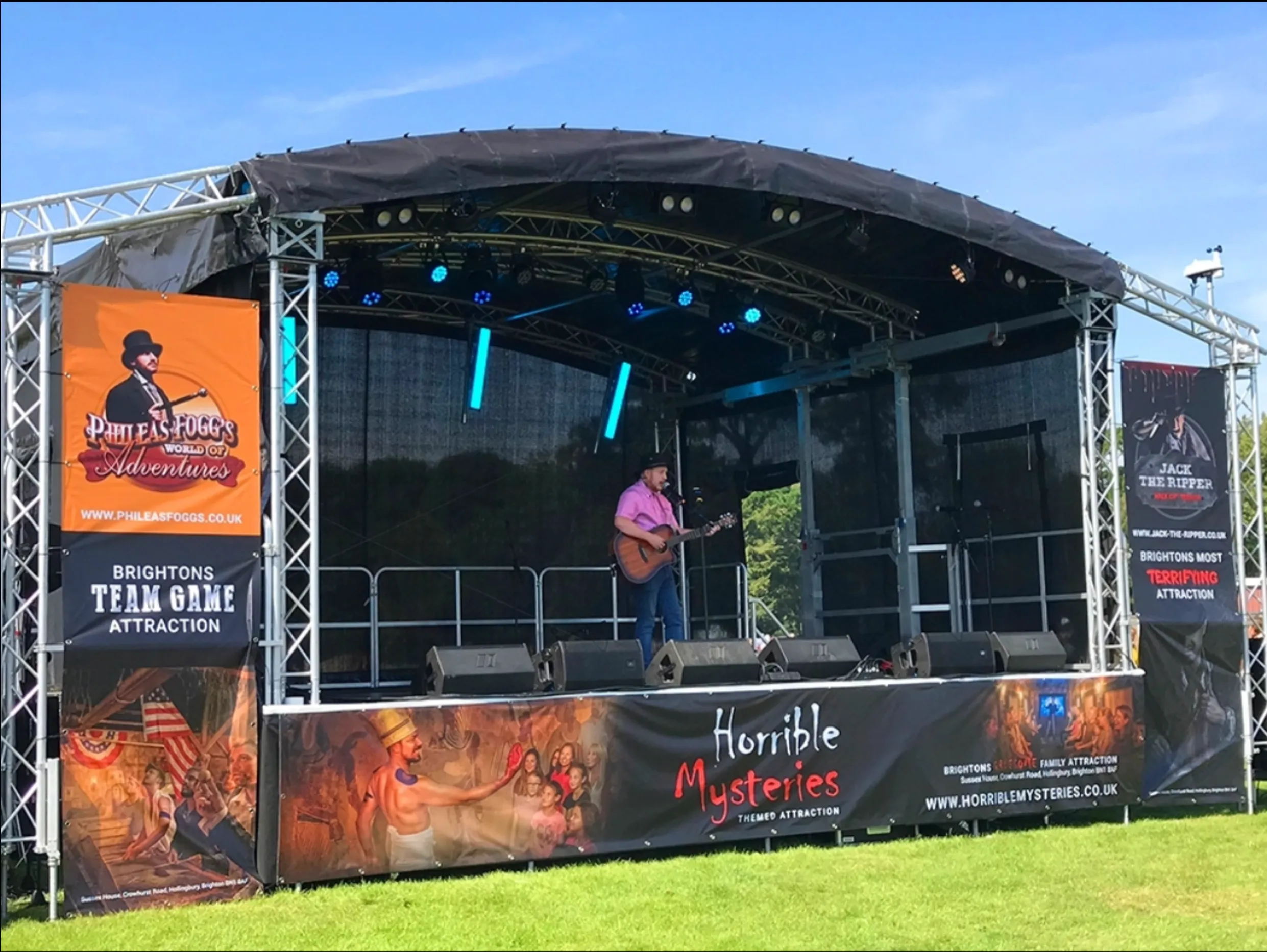 Musician performing on a covered mobile stage on grass with a lighting rig and PA speakers