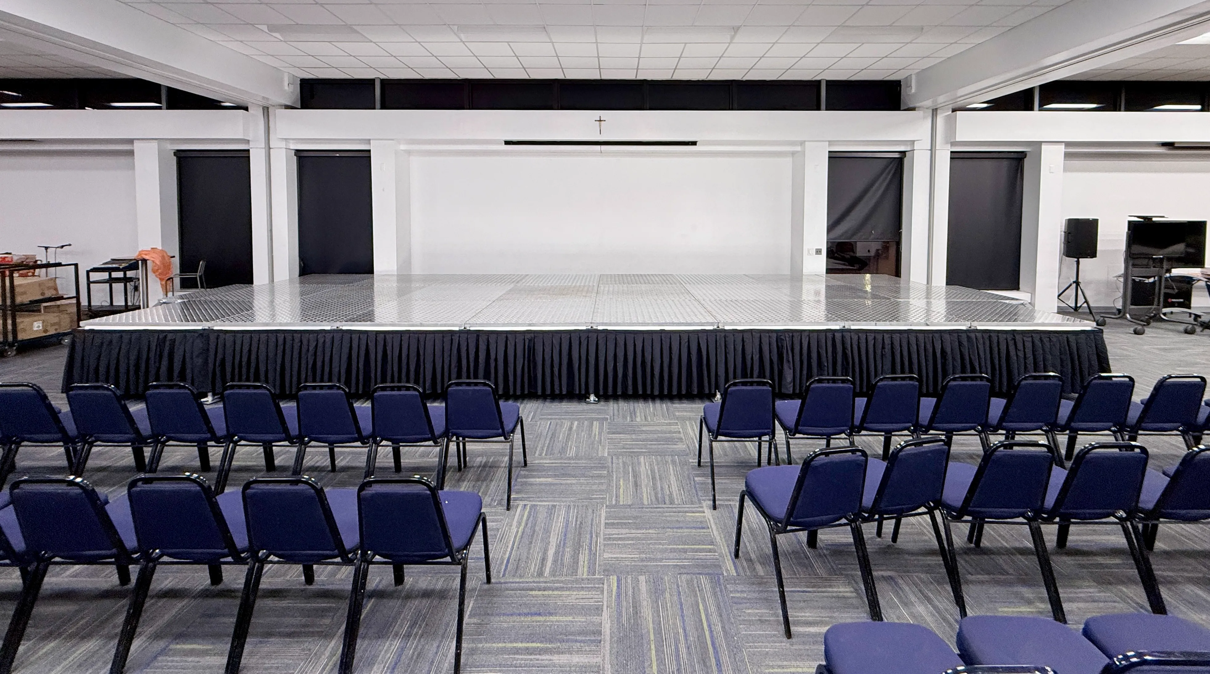 Rows of blue chairs facing a low portable stage with black skirting and a projection screen