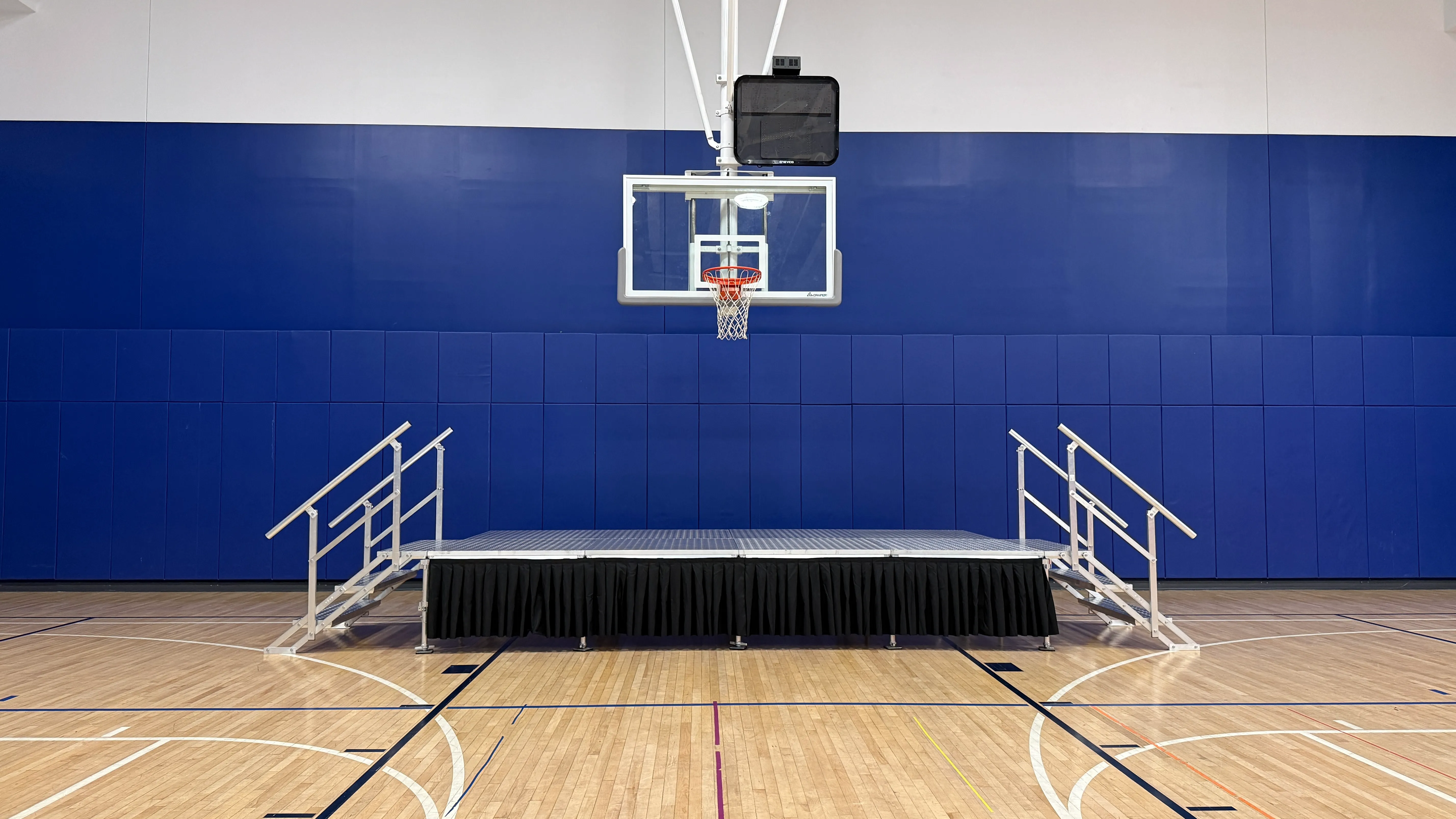 Portable stage with black skirting and handrail steps on both sides beneath a basketball hoop