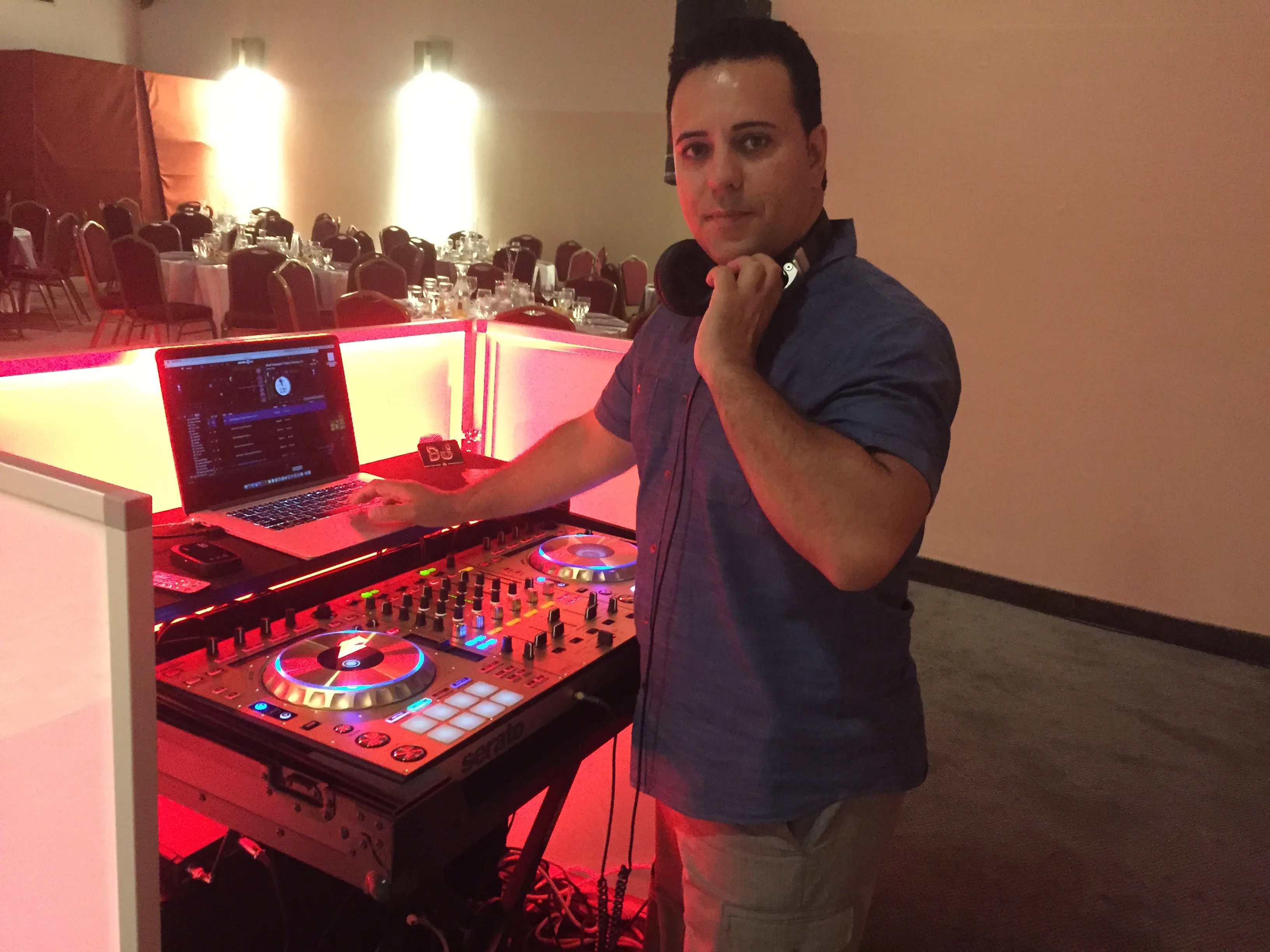 DJ standing at a white lit booth with a controller and laptop in a banquet room set for a wedding