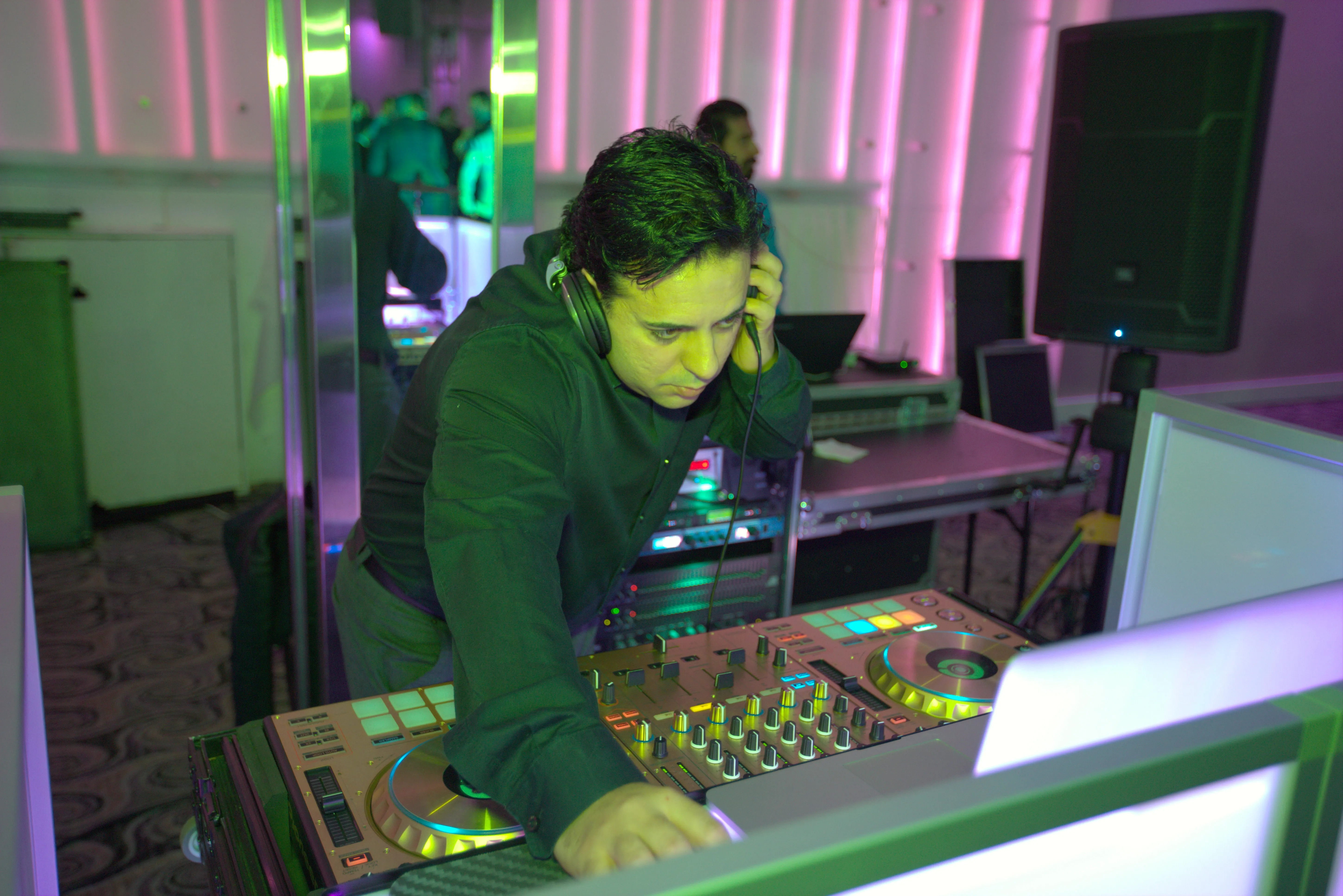 DJ wearing headphones working a controller at a lit booth with a powered speaker on a stand behind